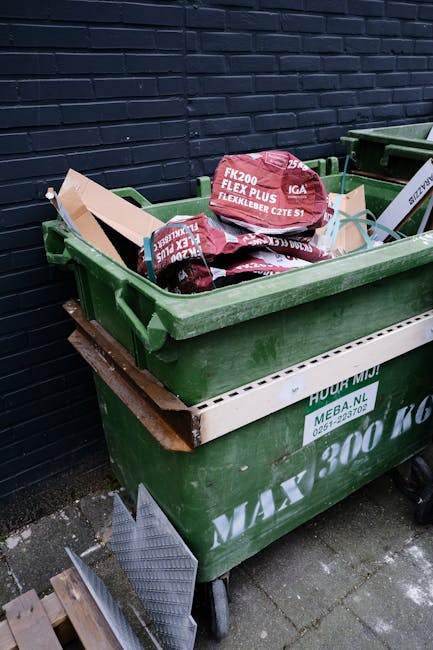 A row of large, upright rubbish bins placed outdoors along a narrow paved pathway beside a brick wall. The bins are made of durable plastic with textured lids, each featuring a distinct colour: dark g