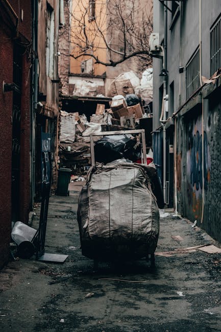 A white commercial van parked on a narrow city street beside a multi-storey building with a grey stone facade. The van's rear doors are open, revealing a large assortment of waste materials including 